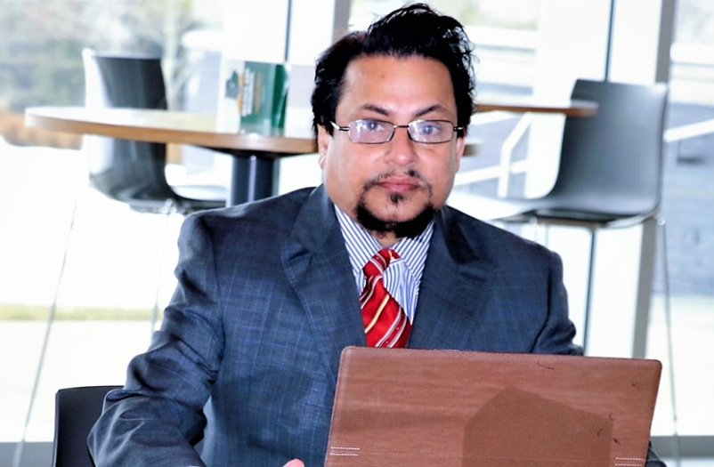 Man with short black hair wearing glasses, dressed in a navy blue suit with a red tie, on his computer sitting in a library.